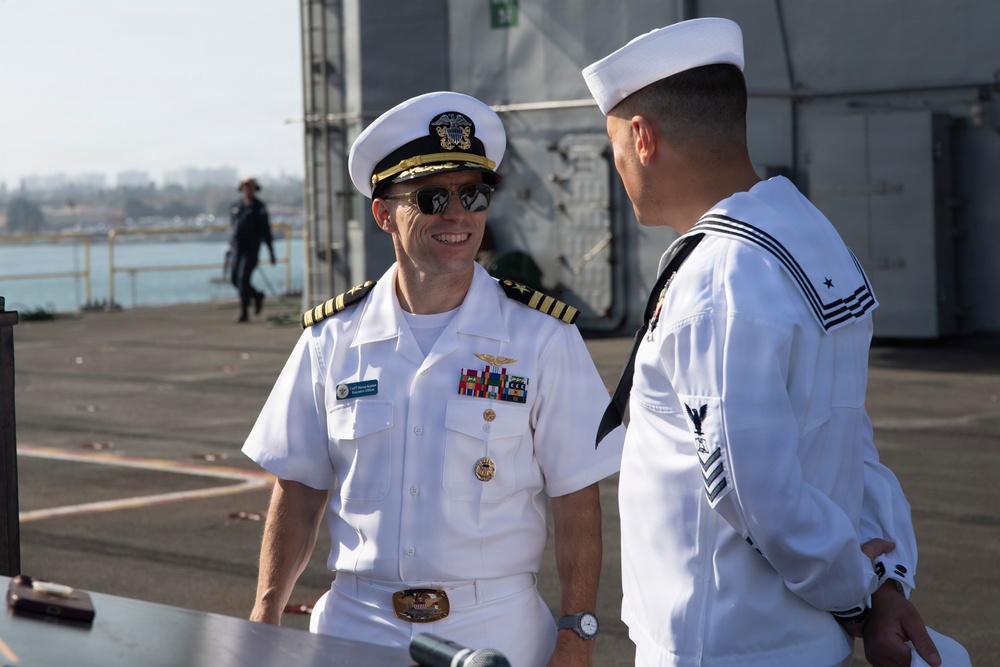 Two U.S. Navy personnel are engaged in conversation on what appears to be the deck of a naval vessel at a port. The officer on the left wears a Captain's rank (four gold stripes) with aviator wings, multiple service ribbons, and a white dress uniform, while the enlisted sailor on the right wears a Dress White uniform with Petty Officer insignia. The setting appears to be a formal or ceremonial occasion based on their dress uniforms.