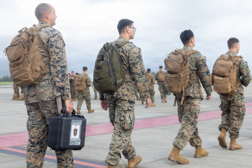 A group of military personnel in digital camouflage uniforms are walking across what appears to be an airfield tarmac. The soldiers are carrying tactical backpacks and at least one hard-shell equipment case labeled 'BOX FOUR'. The scene suggests a deployment, arrival, or movement operation at a military airfield.