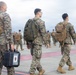 A group of military personnel in digital camouflage uniforms are walking across what appears to be an airfield tarmac. The soldiers are carrying tactical backpacks and at least one hard-shell equipment case labeled 'BOX FOUR'. The scene suggests a deployment, arrival, or movement operation at a military airfield.