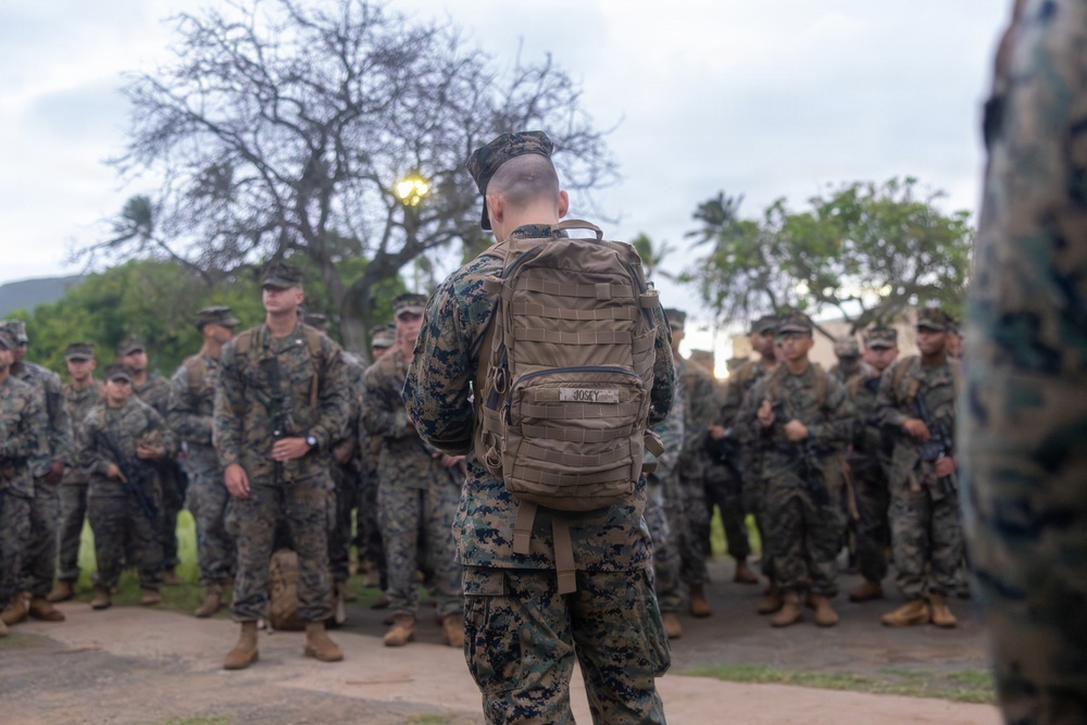 A military formation scene showing a Marine with a tactical backpack labeled 'JOSEY' addressing a large group of fellow Marines in digital camouflage (MARPAT) uniforms. The setting appears to be an outdoor area with trees and a lamp post in the background, likely on a military installation. The Marines are standing in formation holding rifles while being briefed or addressed.