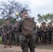 A military formation scene showing a Marine with a tactical backpack labeled 'JOSEY' addressing a large group of fellow Marines in digital camouflage (MARPAT) uniforms. The setting appears to be an outdoor area with trees and a lamp post in the background, likely on a military installation. The Marines are standing in formation holding rifles while being briefed or addressed.