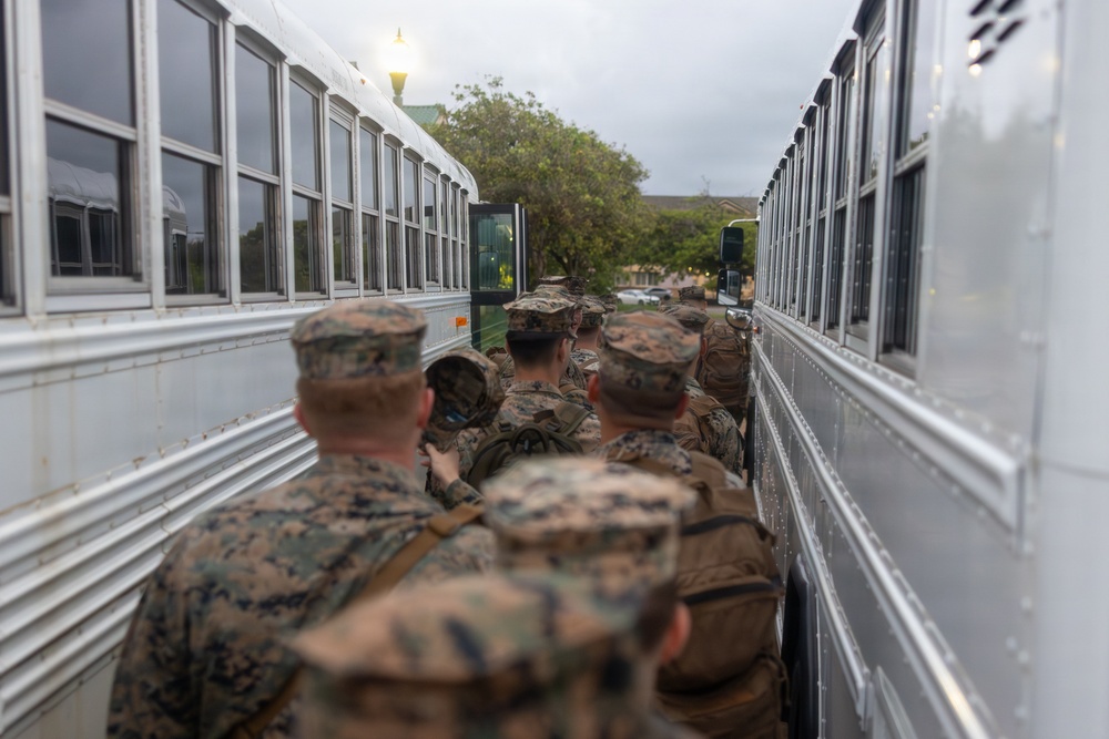 A group of military personnel in camouflage uniforms are boarding or queuing between two large white buses. The soldiers are carrying backpacks and appear to be preparing for transport, likely as part of a deployment or training exercise. The scene takes place outdoors in overcast conditions near what appears to be a military installation.