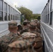 A group of military personnel in camouflage uniforms are boarding or queuing between two large white buses. The soldiers are carrying backpacks and appear to be preparing for transport, likely as part of a deployment or training exercise. The scene takes place outdoors in overcast conditions near what appears to be a military installation.