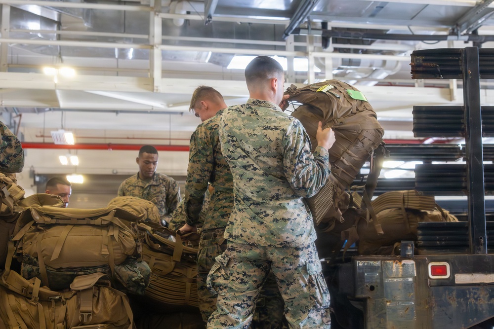 Military personnel in digital camouflage uniforms are loading or unloading large tactical backpacks and gear onto a flatbed truck inside what appears to be a warehouse or hangar facility. Multiple service members are actively handling heavy equipment bags, with one individual hoisting a large pack onto the truck bed. The setting suggests a deployment, logistics operation, or equipment staging activity.
