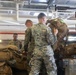 Military personnel in digital camouflage uniforms are loading or unloading large tactical backpacks and gear onto a flatbed truck inside what appears to be a warehouse or hangar facility. Multiple service members are actively handling heavy equipment bags, with one individual hoisting a large pack onto the truck bed. The setting suggests a deployment, logistics operation, or equipment staging activity.