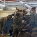 Several military personnel in MARPAT (Marine digital camouflage) uniforms are handling and organizing tactical gear and equipment inside an industrial facility. The individuals appear to be loading, stacking, or inspecting body armor, tactical vests, and backpacks. The setting appears to be a warehouse or equipment staging area.