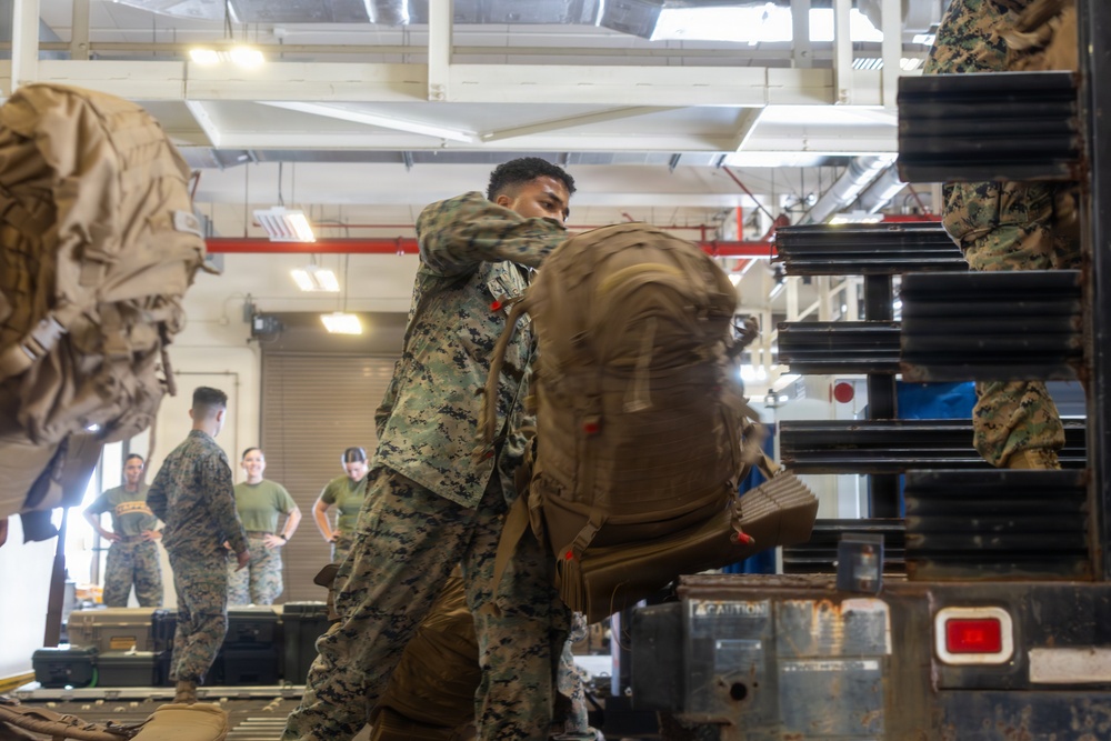 Military personnel in MARPAT camouflage uniforms are loading large tactical backpacks onto what appears to be a conveyor or loading ramp inside a warehouse or hangar facility. The central figure is actively hoisting a large tan/coyote-colored rucksack onto the equipment, while other service members observe or assist in the background. The scene depicts a deployment or logistics operation involving the movement of military gear.