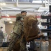 Military personnel in MARPAT camouflage uniforms are loading large tactical backpacks onto what appears to be a conveyor or loading ramp inside a warehouse or hangar facility. The central figure is actively hoisting a large tan/coyote-colored rucksack onto the equipment, while other service members observe or assist in the background. The scene depicts a deployment or logistics operation involving the movement of military gear.