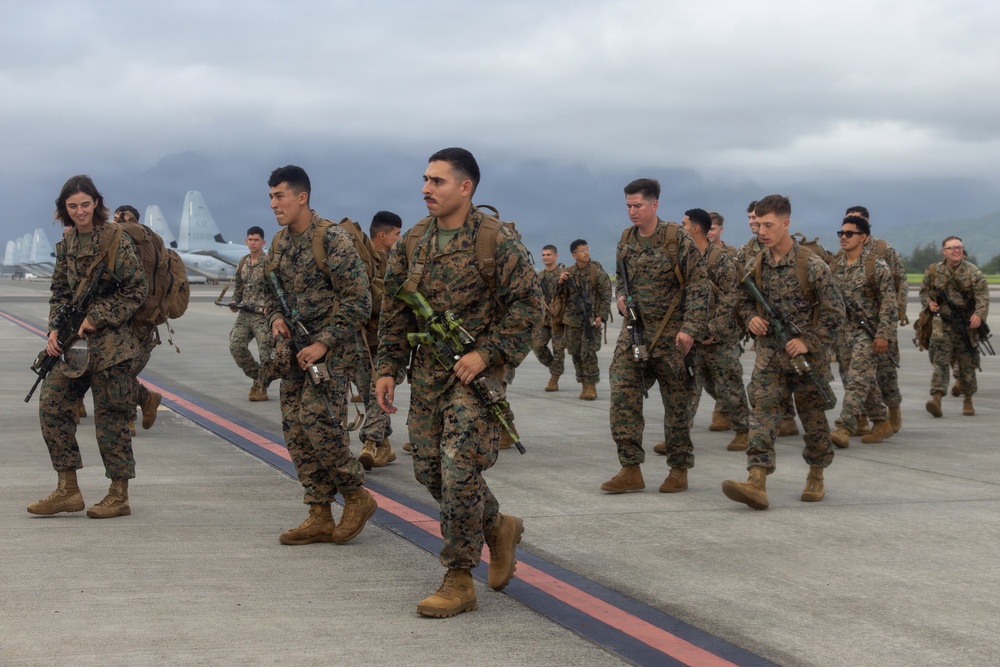 A large group of uniformed military personnel, appearing to be U.S. Marines based on their MARPAT camouflage uniforms, are marching across an airfield tarmac while carrying rifles and full combat gear. The scene appears to take place at a military airbase, with aircraft visible in the background and mountainous terrain obscured by overcast skies. Both male and female service members are present in the formation.