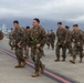 A large group of uniformed military personnel, appearing to be U.S. Marines based on their MARPAT camouflage uniforms, are marching across an airfield tarmac while carrying rifles and full combat gear. The scene appears to take place at a military airbase, with aircraft visible in the background and mountainous terrain obscured by overcast skies. Both male and female service members are present in the formation.