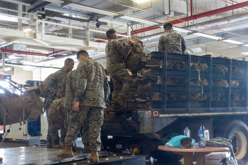 Several military personnel in woodland digital camouflage uniforms are loading or unloading tactical gear and backpacks onto/from a large flatbed military truck inside what appears to be a vehicle bay or warehouse facility. The truck bed is heavily loaded with tan/coyote-colored military rucksacks and equipment. The indoor setting features exposed piping and industrial ceiling infrastructure.