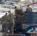 Several military personnel in woodland digital camouflage uniforms are loading or unloading tactical gear and backpacks onto/from a large flatbed military truck inside what appears to be a vehicle bay or warehouse facility. The truck bed is heavily loaded with tan/coyote-colored military rucksacks and equipment. The indoor setting features exposed piping and industrial ceiling infrastructure.
