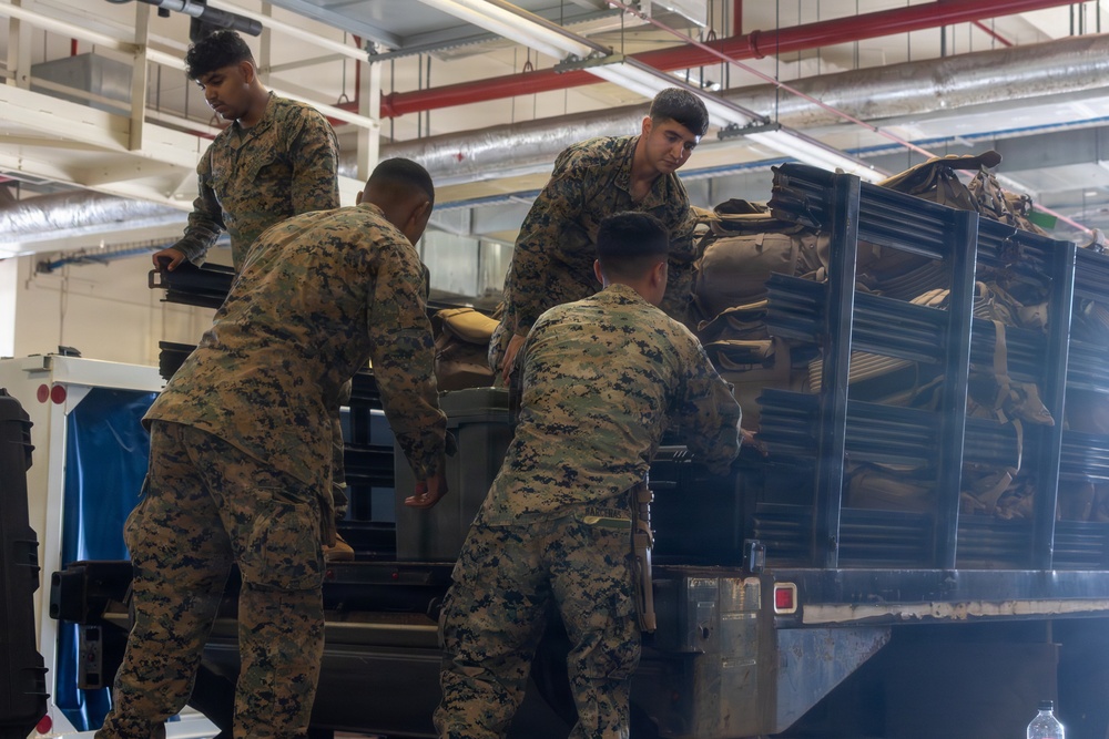 Four military personnel in MARPAT (Marine pattern) camouflage uniforms are loading or unloading equipment onto a flatbed truck inside what appears to be a warehouse or hangar facility. The soldiers are handling various military gear including containers, cases, and packed equipment. The scene depicts a logistics or deployment preparation operation.
