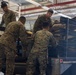 Four military personnel in MARPAT (Marine pattern) camouflage uniforms are loading or unloading equipment onto a flatbed truck inside what appears to be a warehouse or hangar facility. The soldiers are handling various military gear including containers, cases, and packed equipment. The scene depicts a logistics or deployment preparation operation.