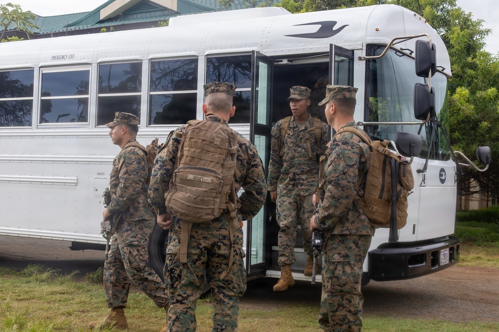 A group of military personnel in MARPAT (Marine Pattern) camouflage uniforms are boarding or gathering around a white bus. The soldiers are equipped with tactical backpacks and gear, suggesting a deployment, transport, or training exercise. The setting appears to be a military installation with green vegetation and a building visible in the background.