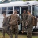 A group of military personnel in MARPAT (Marine Pattern) camouflage uniforms are boarding or gathering around a white bus. The soldiers are equipped with tactical backpacks and gear, suggesting a deployment, transport, or training exercise. The setting appears to be a military installation with green vegetation and a building visible in the background.