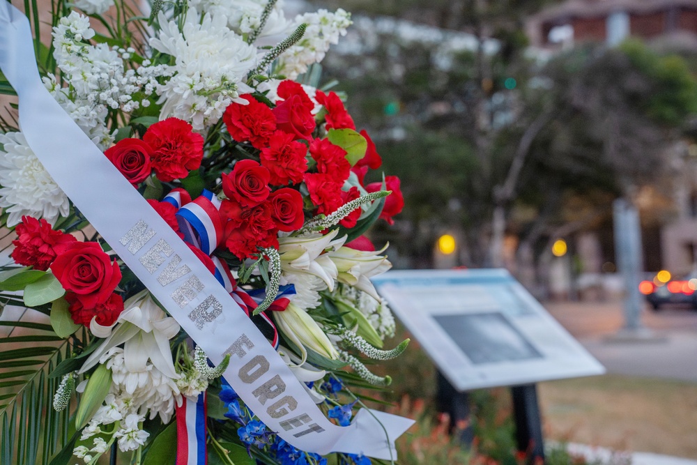 A memorial wreath featuring red, white, and blue flowers is displayed at what appears to be a commemorative ceremony. The wreath bears a white sash with the visible text 'NEVER FORGET,' indicating a patriotic or military remembrance event. An informational placard or memorial sign is visible in the blurred background.