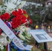 A memorial wreath featuring red, white, and blue flowers is displayed at what appears to be a commemorative ceremony. The wreath bears a white sash with the visible text 'NEVER FORGET,' indicating a patriotic or military remembrance event. An informational placard or memorial sign is visible in the blurred background.