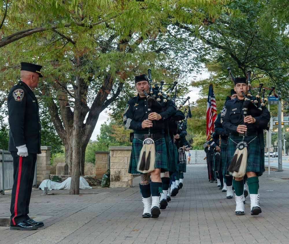 A law enforcement pipe and drum band marches in formal procession along a tree-lined brick walkway. The bagpipers are dressed in dark uniforms with tartan kilts, white spats, and Glengarry caps, while a senior officer in dress uniform stands at attention to the left. An American flag is visible being carried in the procession behind the lead pipers.