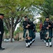 A law enforcement pipe and drum band marches in formal procession along a tree-lined brick walkway. The bagpipers are dressed in dark uniforms with tartan kilts, white spats, and Glengarry caps, while a senior officer in dress uniform stands at attention to the left. An American flag is visible being carried in the procession behind the lead pipers.