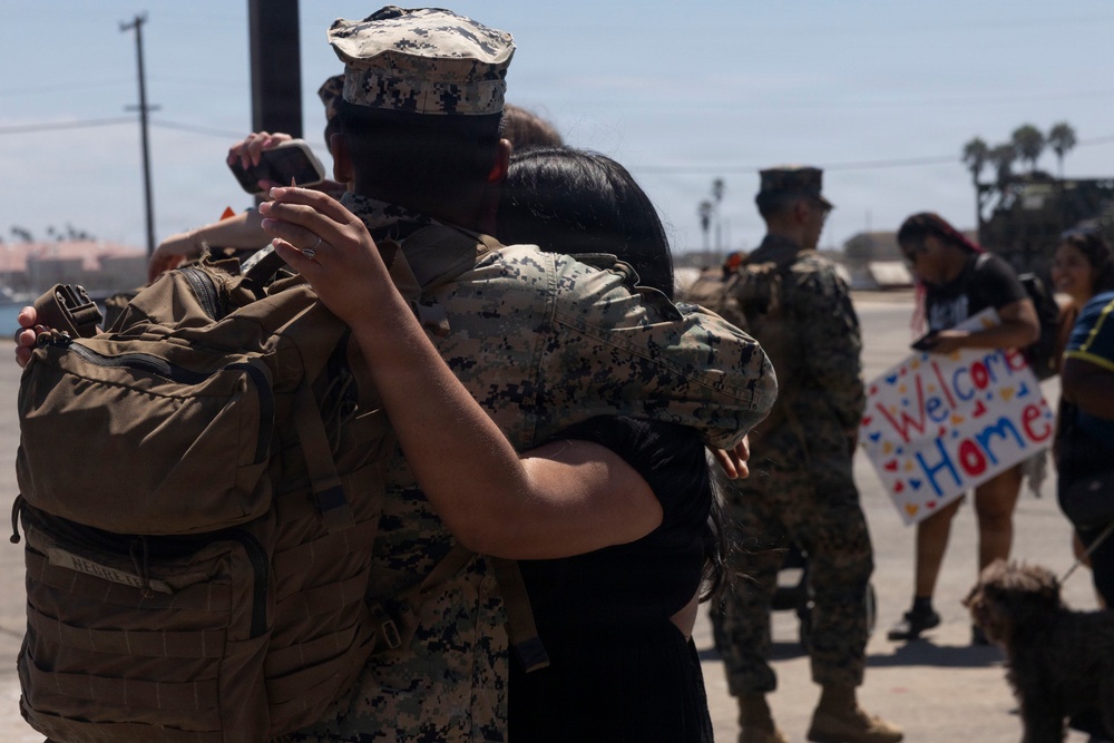 A military homecoming scene showing a uniformed service member embracing a civilian, likely a family member or loved one. The setting appears to be an outdoor tarmac or base area, with other military personnel and civilians visible in the background. A handmade 'Welcome Home' sign is visible in the background, confirming this is a return-from-deployment event.