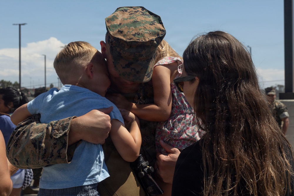 A U.S. Marine in woodland MARPAT camouflage uniform is embracing two young children simultaneously, with a woman standing closely beside them. The scene appears to be a military homecoming or farewell event, taking place outdoors on a military installation. The emotional reunion captures a deeply personal family moment amid what appears to be a larger gathering of military personnel and families.