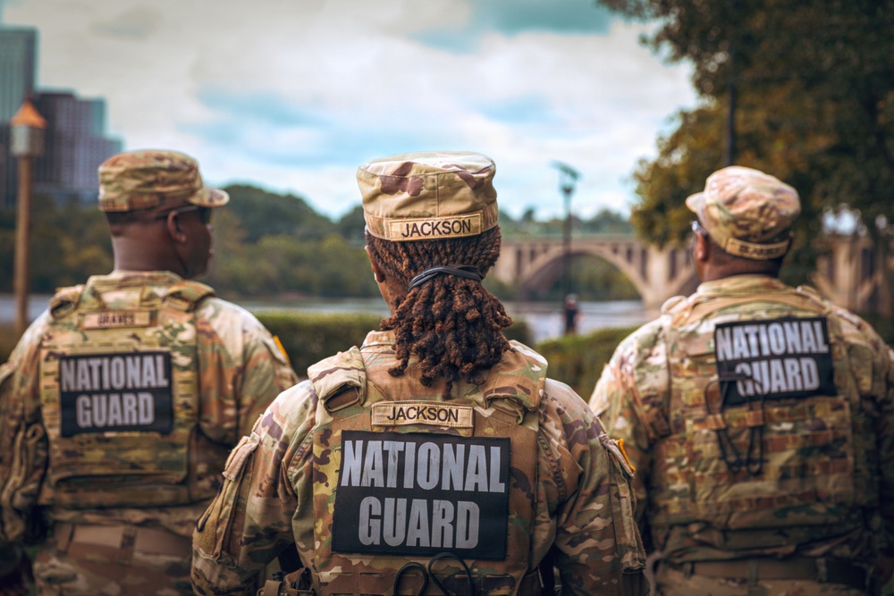 Three National Guard soldiers are photographed from behind, wearing multicam/OCP camouflage uniforms with 'NATIONAL GUARD' patches on their backs. The center soldier has the name tape 'JACKSON' visible on both their cap and uniform. They appear to be standing near a waterway with a stone arch bridge visible in the background.