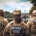 Three National Guard soldiers are photographed from behind, wearing multicam/OCP camouflage uniforms with 'NATIONAL GUARD' patches on their backs. The center soldier has the name tape 'JACKSON' visible on both their cap and uniform. They appear to be standing near a waterway with a stone arch bridge visible in the background.