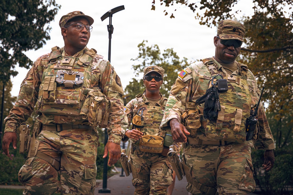 Three U.S. Army soldiers in OCP (Operational Camouflage Pattern) uniforms are walking together in what appears to be an outdoor urban or park setting. All three are wearing tactical plate carriers and combat gear, with name tapes visible on at least two individuals ('BLAKELY' on the left and 'GRAY' on the right). The soldier on the right is carrying a tactical radio, and all appear to be in an active patrol or movement posture.