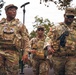 Three U.S. Army soldiers in OCP (Operational Camouflage Pattern) uniforms are walking together in what appears to be an outdoor urban or park setting. All three are wearing tactical plate carriers and combat gear, with name tapes visible on at least two individuals ('BLAKELY' on the left and 'GRAY' on the right). The soldier on the right is carrying a tactical radio, and all appear to be in an active patrol or movement posture.