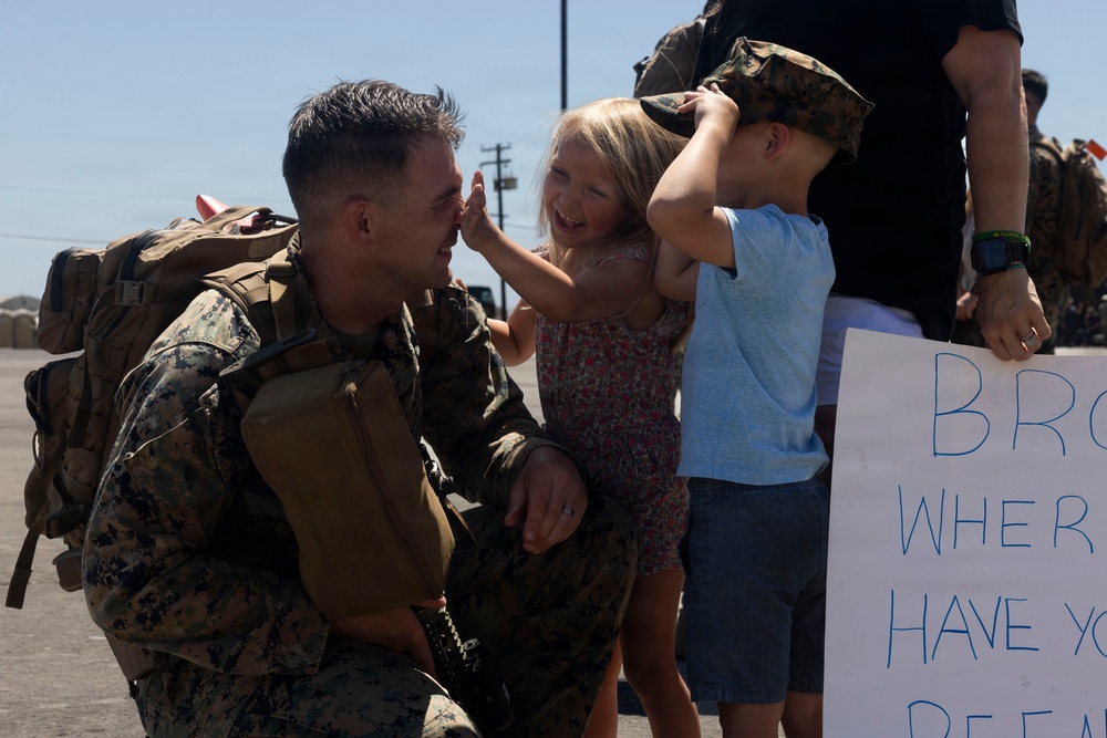 A U.S. Marine in full combat uniform and gear kneels down to greet two young children during what appears to be a military homecoming event. A young girl is laughing and touching the Marine's face while a boy tries on the Marine's camouflage cover (hat). A partially visible sign in the background reads 'BRO WHERE HAVE YO BE...' suggesting a family reunion.