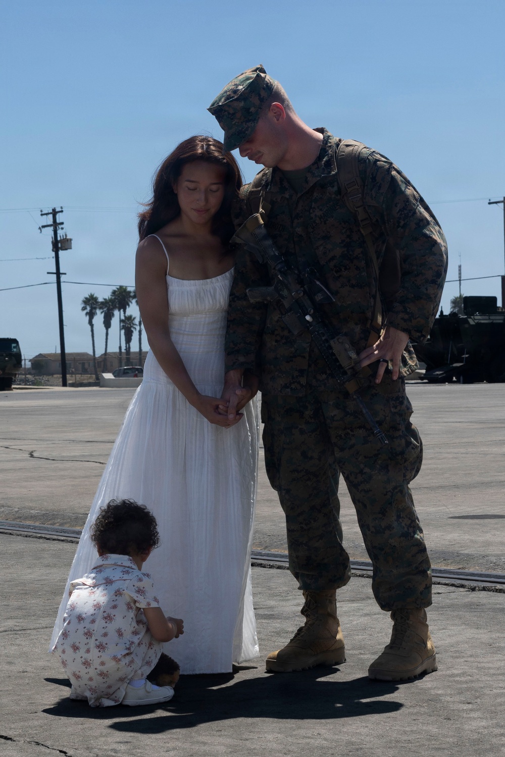 A U.S. Marine in full combat uniform and gear stands with a woman in a white dress and a small toddler on a military base tarmac. The adults are holding hands and leaning their heads together in an intimate moment, while the young child crouches at their feet. The setting and gear suggest a deployment departure or homecoming event.