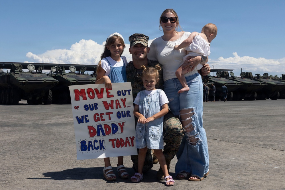 A military service member in camouflage uniform is reuniting with his family at what appears to be a military base or airfield. The family, consisting of a woman and three young children including an infant, holds a handmade sign celebrating his return. Armored military vehicles are visible in the background.