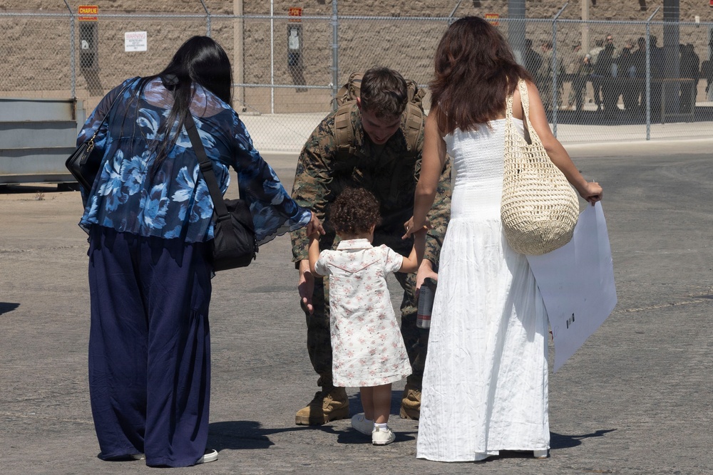 A military service member in camouflage uniform crouches down to interact with a small toddler on what appears to be a military base or installation. Two women flank the service member and child, appearing to participate in what looks like a reunion or farewell moment. The setting is an outdoor paved area with chain-link fencing and military base signage visible in the background.