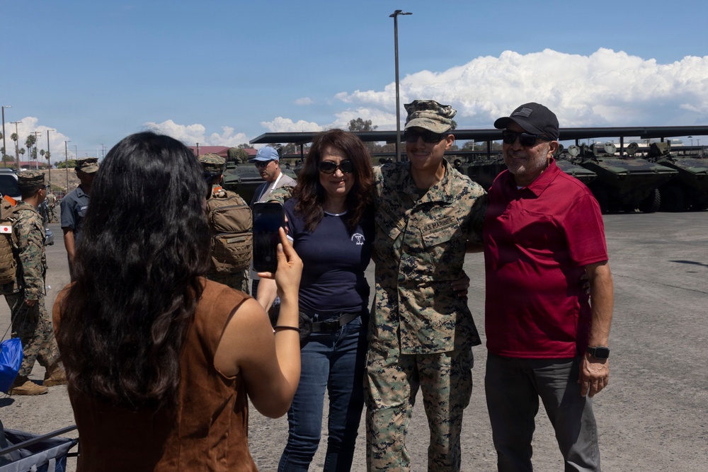 A U.S. Marine in camouflage uniform poses for a photo with two civilians, likely family members, at what appears to be a military open house or base event. A young woman photographs the group using a smartphone. Military vehicles, including armored personnel carriers, and other uniformed personnel are visible in the background.