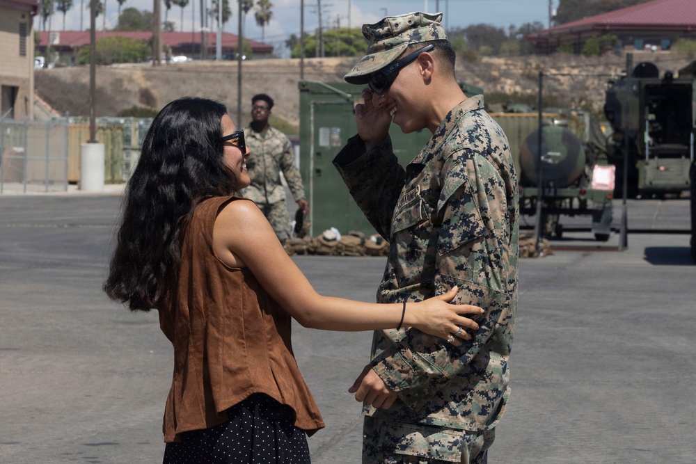 A civilian woman in a brown sleeveless top and sunglasses shares a joyful, intimate moment with a service member in U.S. Marine Corps MARPAT digital camouflage uniform on a military base. The two are smiling and leaning toward each other in what appears to be a reunion or farewell encounter. The setting is an outdoor paved area of a military installation with military vehicles and equipment visible in the background.