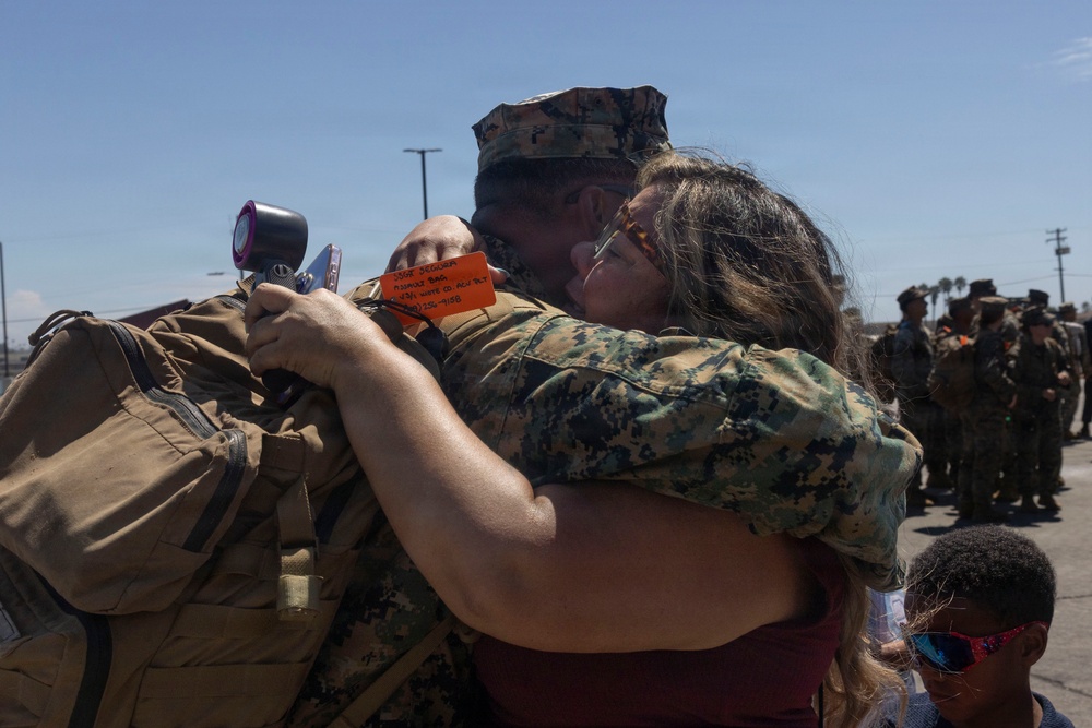 A U.S. Marine in digital camouflage uniform and cover (cap) is embracing a civilian woman in an emotional hug, likely during a deployment departure or homecoming event. The Marine is wearing a large backpack with an orange identification tag attached. Other Marines in formation are visible in the background, and a young child is partially visible in the lower right corner.