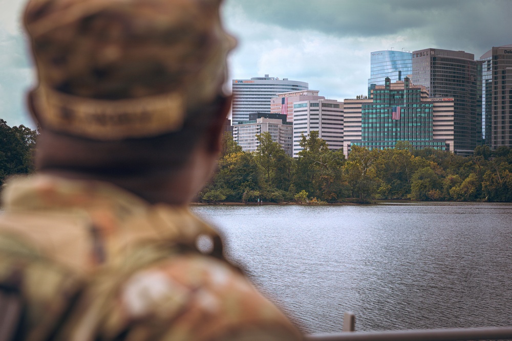 A military service member in camouflage uniform and patrol cap is seen from behind, looking across a body of water toward an urban skyline. The cityscape features multiple mid-to-high-rise office buildings, at least two of which display American flags. The scene appears to be taken from a waterfront vantage point under overcast skies.