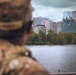 A military service member in camouflage uniform and patrol cap is seen from behind, looking across a body of water toward an urban skyline. The cityscape features multiple mid-to-high-rise office buildings, at least two of which display American flags. The scene appears to be taken from a waterfront vantage point under overcast skies.