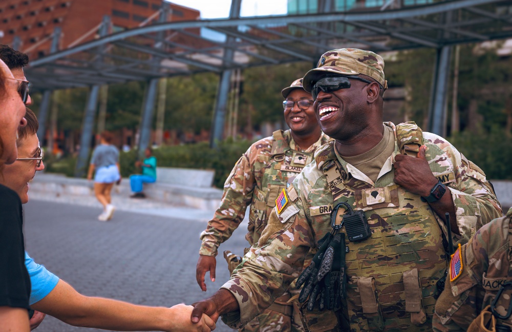 Two U.S. Army soldiers in OCP (Operational Camouflage Pattern) uniforms are interacting with civilians in what appears to be an urban public space. The foreground soldier, whose name tape reads 'GRA-' (partially visible), is laughing and shaking hands with a civilian while wearing tactical gear including a plate carrier and radio. The interaction appears friendly and community-oriented.