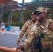 Two U.S. Army soldiers in OCP (Operational Camouflage Pattern) uniforms are interacting with civilians in what appears to be an urban public space. The foreground soldier, whose name tape reads 'GRA-' (partially visible), is laughing and shaking hands with a civilian while wearing tactical gear including a plate carrier and radio. The interaction appears friendly and community-oriented.