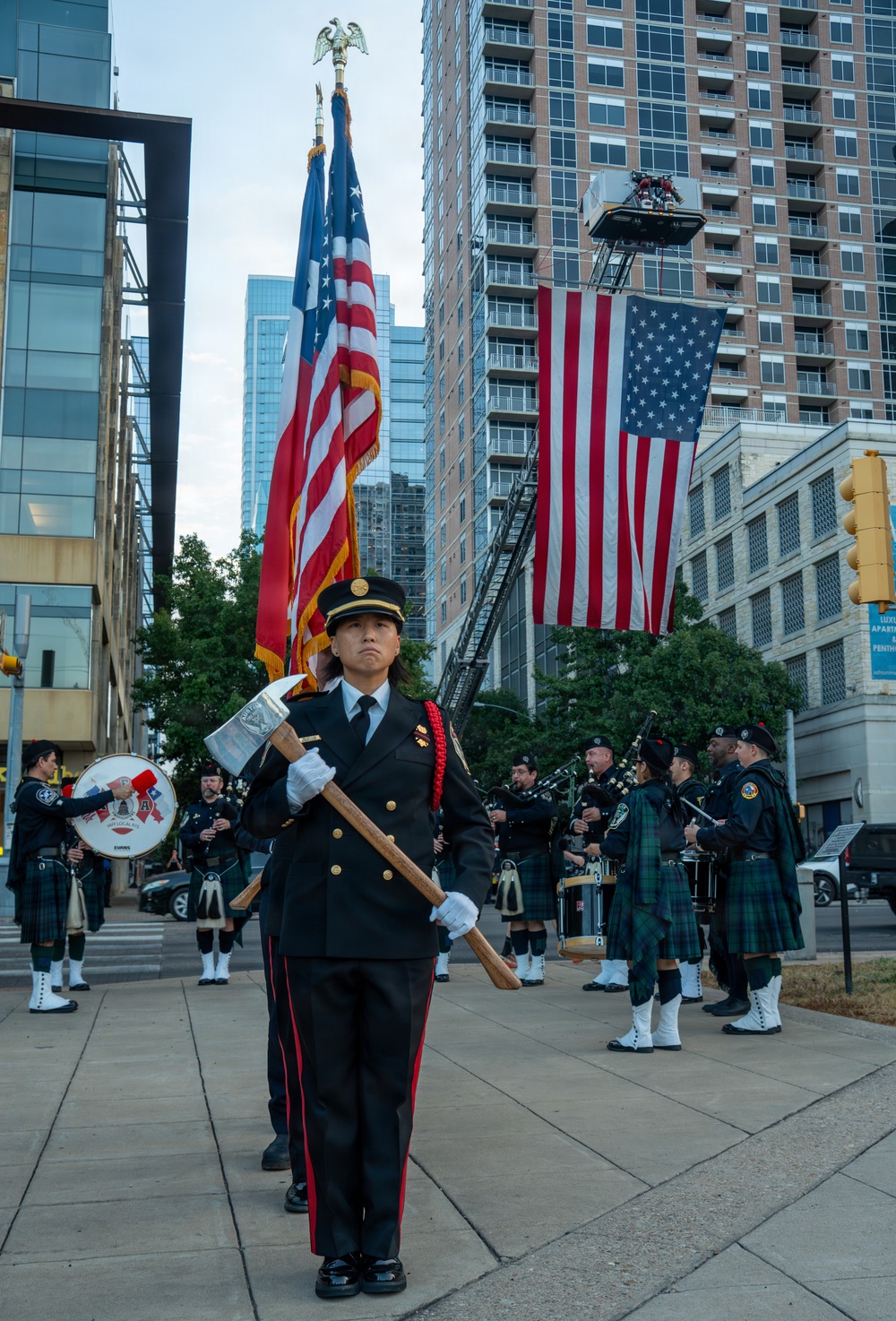 A formal firefighter honor guard ceremony is taking place in an urban setting, with a female firefighter in dress uniform prominently carrying an axe at the forefront. Behind her, a pipe and drum band in kilts performs, while two large American flags are displayed — one carried on a ceremonial pole and one suspended from a fire truck aerial ladder. The setting and imagery are consistent with a 9/11 memorial or first responder commemorative event.