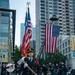 A formal firefighter honor guard ceremony is taking place in an urban setting, with a female firefighter in dress uniform prominently carrying an axe at the forefront. Behind her, a pipe and drum band in kilts performs, while two large American flags are displayed — one carried on a ceremonial pole and one suspended from a fire truck aerial ladder. The setting and imagery are consistent with a 9/11 memorial or first responder commemorative event.