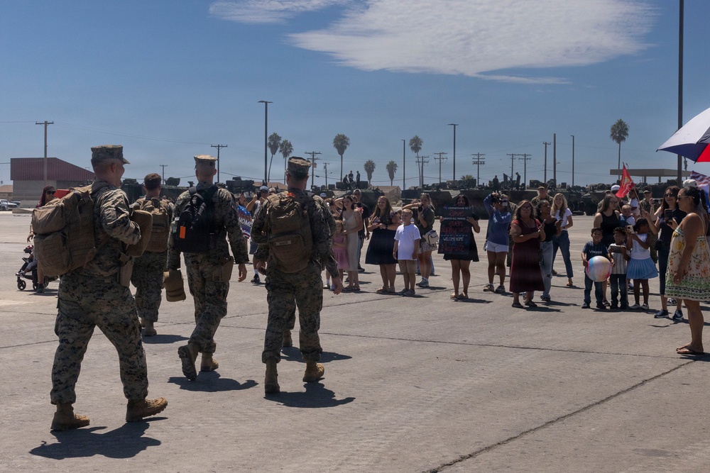 A military homecoming or welcome ceremony is taking place on a large paved area, likely a military base. Three U.S. Marines in camouflage uniforms with backpacks are walking toward a crowd of civilians, including families with children, who are holding welcome signs. Military vehicles are visible in the background, and the setting appears to be a sunny day in a coastal or Southern California environment based on the palm trees.