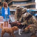Two U.S. Army soldiers in MultiCam (OCP) uniforms crouch down to pet two dogs — one brown-and-white and one golden/auburn — held on leashes by a smiling woman in a blue cardigan and denim shorts. The interaction takes place on a brick plaza in front of what appears to be a waterfront commercial or dining area. The scene conveys a lighthearted, candid moment between military personnel and a civilian dog owner.