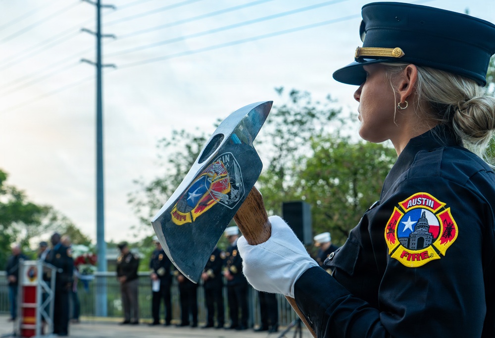 A female Austin Fire Department firefighter in dress uniform holds a ceremonial fire axe bearing the Austin Fire Department emblem during what appears to be a formal memorial or honor ceremony. She is wearing white gloves and a dress cap with gold insignia, standing in profile view. In the background, a group of uniformed personnel and civilians are gathered near a podium, suggesting an official commemorative event.