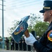 A female Austin Fire Department firefighter in dress uniform holds a ceremonial fire axe bearing the Austin Fire Department emblem during what appears to be a formal memorial or honor ceremony. She is wearing white gloves and a dress cap with gold insignia, standing in profile view. In the background, a group of uniformed personnel and civilians are gathered near a podium, suggesting an official commemorative event.