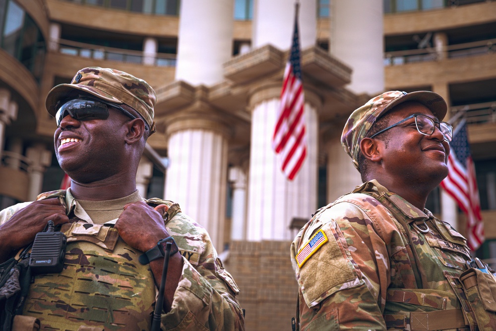 Two U.S. Army soldiers in Operational Camouflage Pattern (OCP) uniforms are standing at attention or on duty in front of a large neoclassical building with prominent white columns. Both soldiers are smiling and appear to be in good spirits, with American flags visible in the background. The setting suggests an official government or civic building, possibly during a ceremonial or security detail.