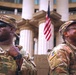 Two U.S. Army soldiers in Operational Camouflage Pattern (OCP) uniforms are standing at attention or on duty in front of a large neoclassical building with prominent white columns. Both soldiers are smiling and appear to be in good spirits, with American flags visible in the background. The setting suggests an official government or civic building, possibly during a ceremonial or security detail.