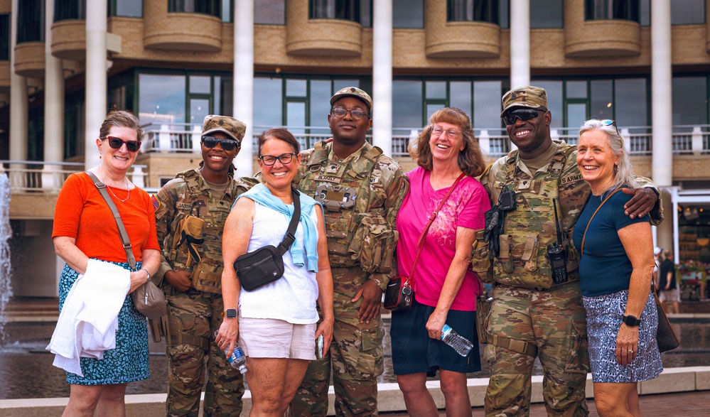 A group of seven people pose together outdoors in front of a large modern building with columns and balconies. Three individuals are wearing U.S. Army camouflage uniforms with tactical gear, while four civilians in casual summer clothing stand among them. The group appears relaxed and smiling, suggesting a friendly, informal interaction.